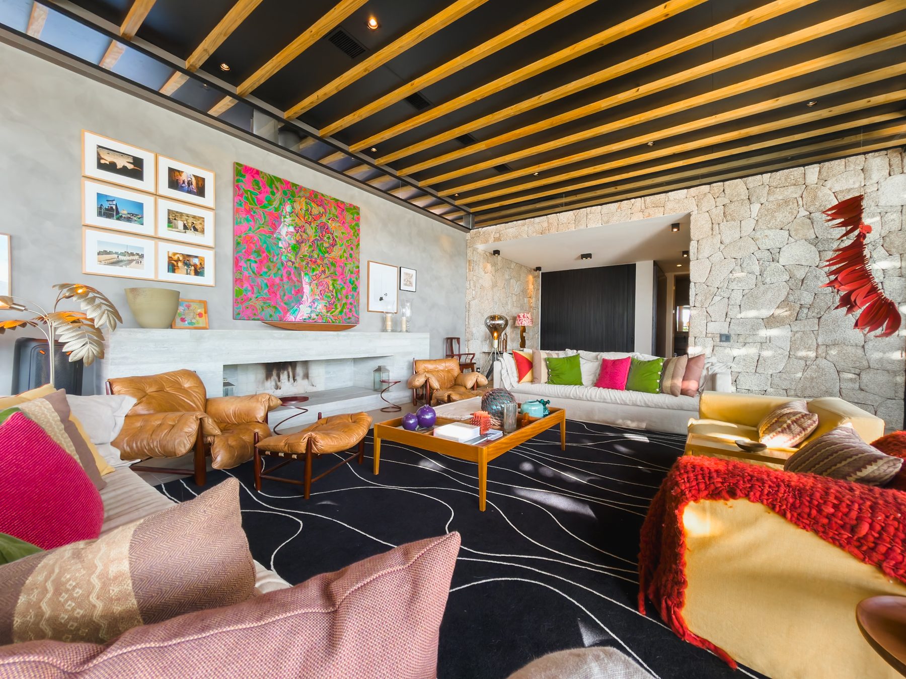 Living amplo sob forro de ripas de madeira, com sofás claros, poltronas de couro e quadro colorido