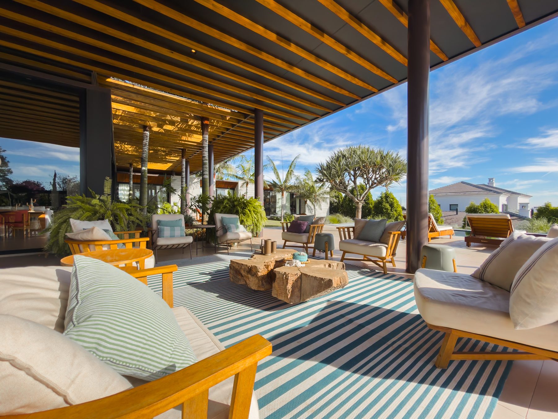 Lounge coberto com sofás de madeira clara sobre tapete listrado, olhando para o jardim