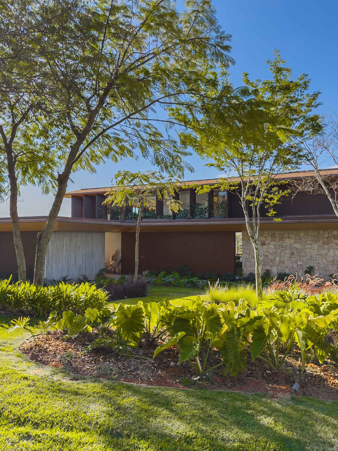 Fachada da casa entre árvores e jardim, com volumes de madeira e pedra