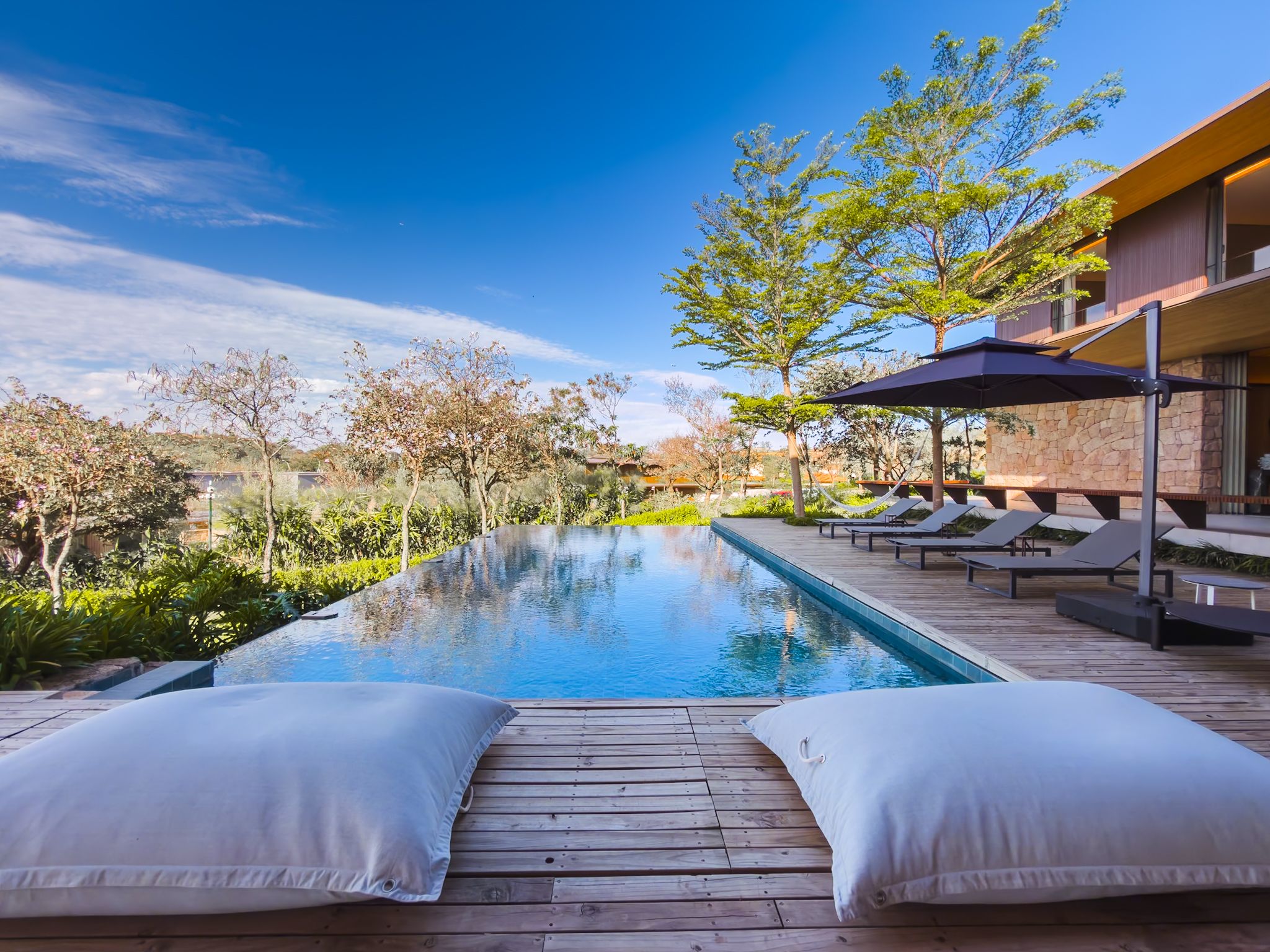 Piscina de borda infinita com almofadas no deck, espreguiçadeiras e o beiral de madeira da casa sob céu azul