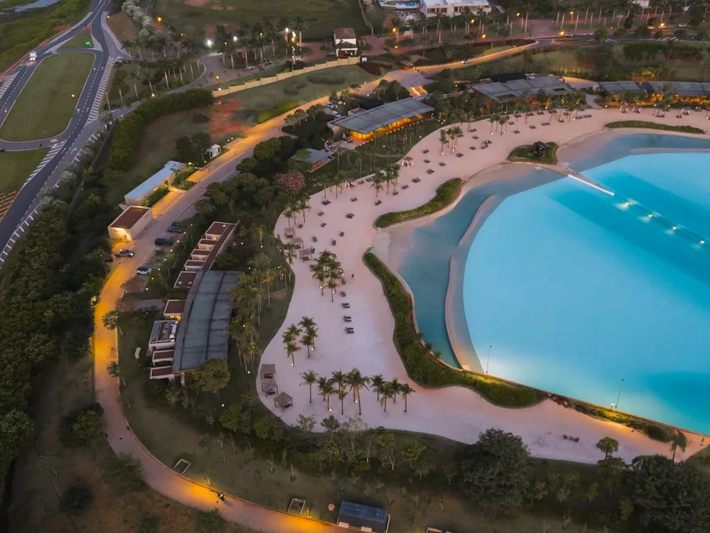 Vista aérea da Praia da Grama ao entardecer, com a lagoa de ondas, a faixa de areia e o beach club iluminado