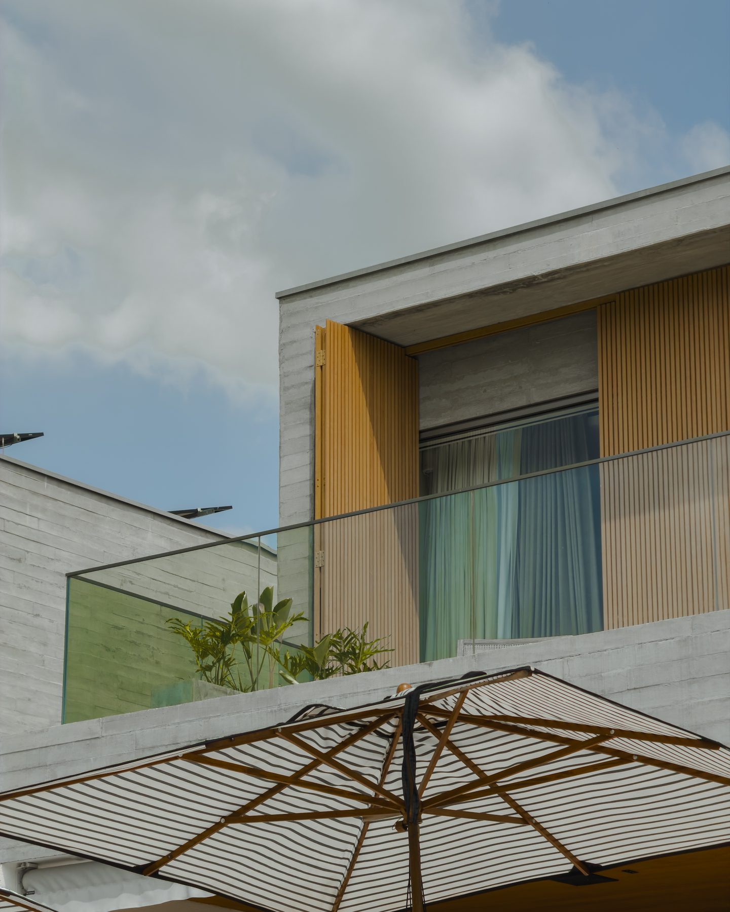 Detalhe da fachada com painéis de madeira, concreto e guarda-corpo de vidro, sobre os guarda-sóis