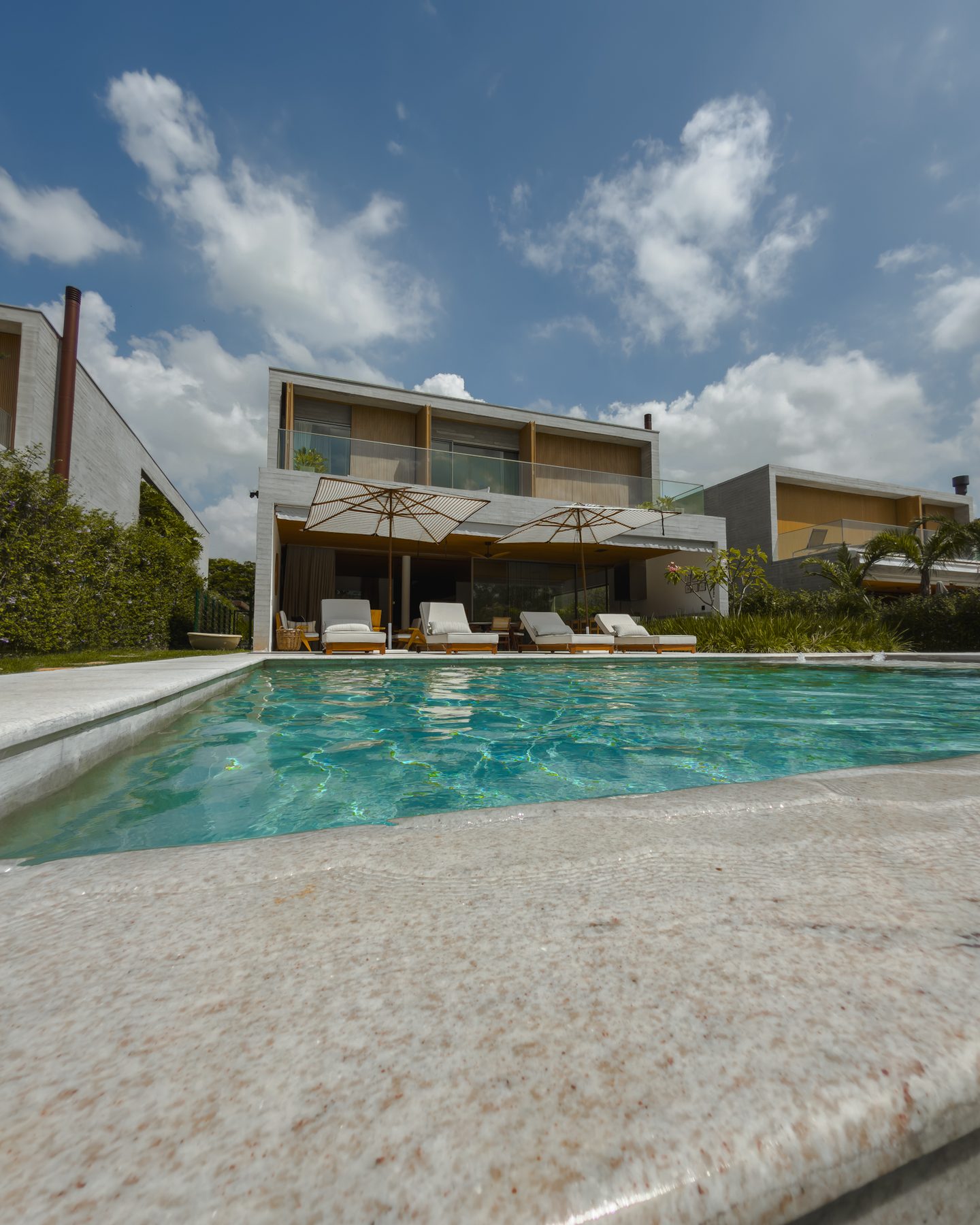 A casa de dois pavimentos vista da borda da piscina, com guarda-sóis e espreguiçadeiras