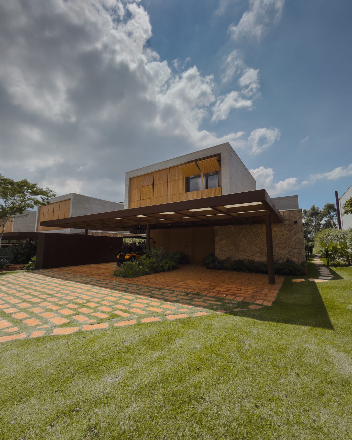 Fachada da casa com volume revestido de madeira, pergolado e piso de tijolos, sob céu com nuvens