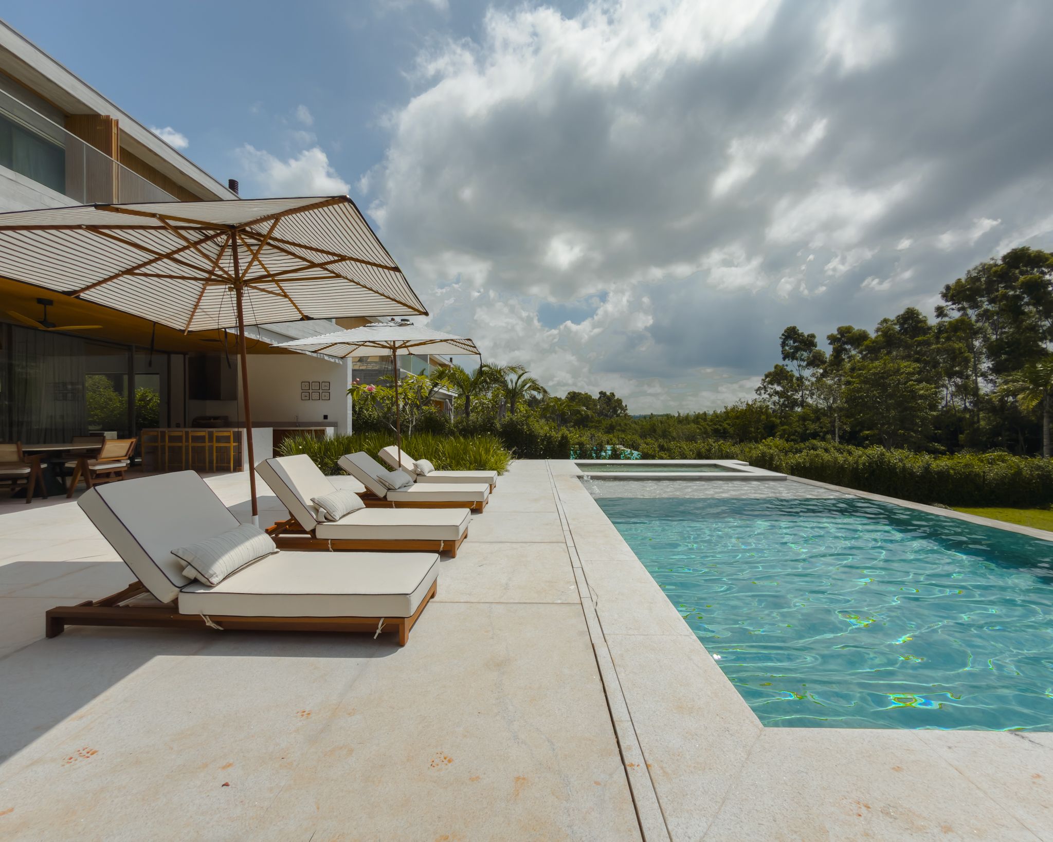 Casa contemporânea de concreto e madeira vista do deck, com espreguiçadeiras, guarda-sóis e a piscina sob céu com nuvens