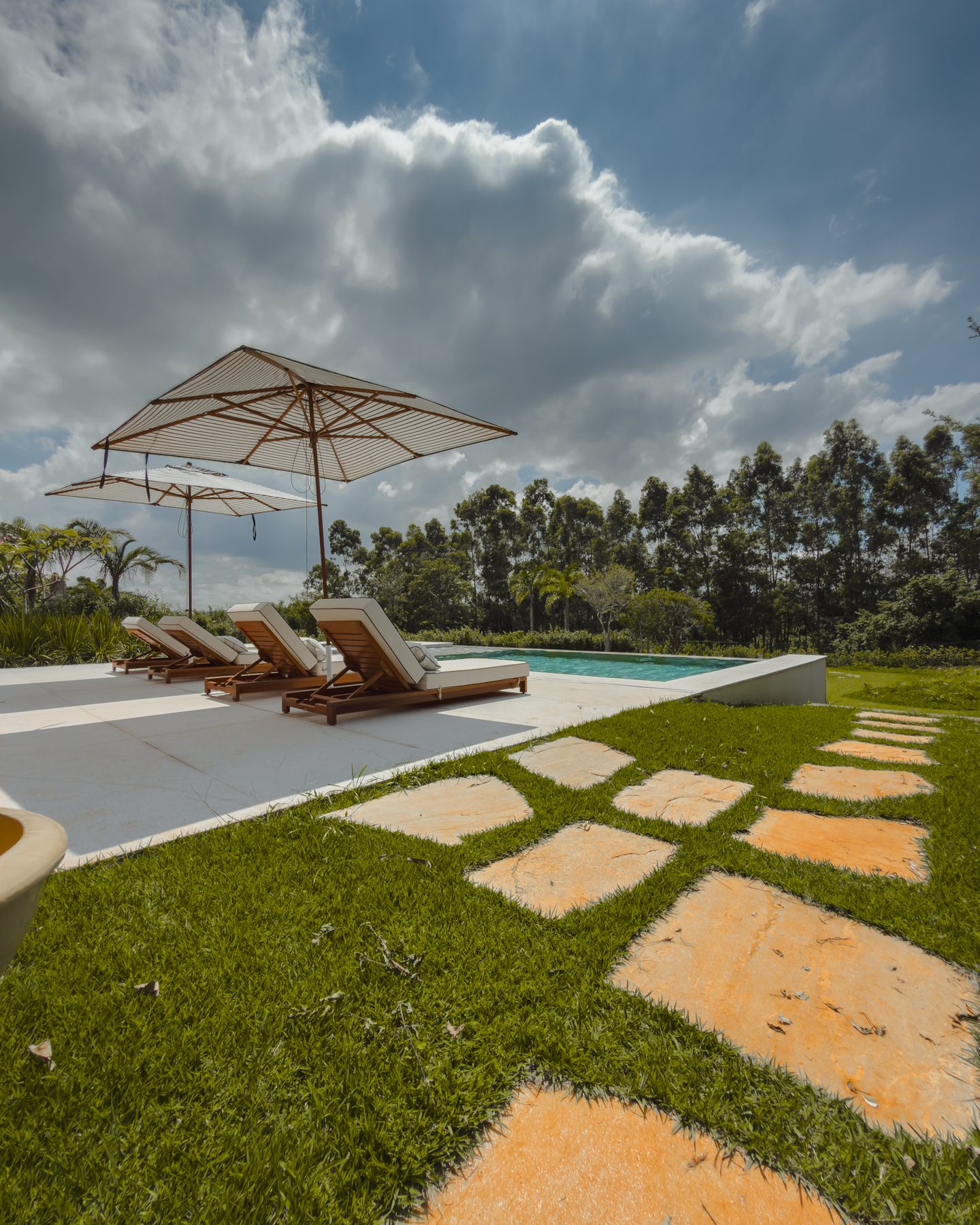 Caminho de pedras no gramado até a piscina, com guarda-sóis e mata ao fundo