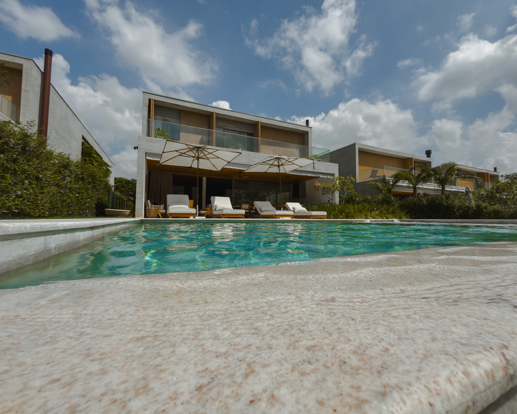 Piscina em primeiro plano, com a casa de concreto e madeira e os guarda-sóis ao fundo