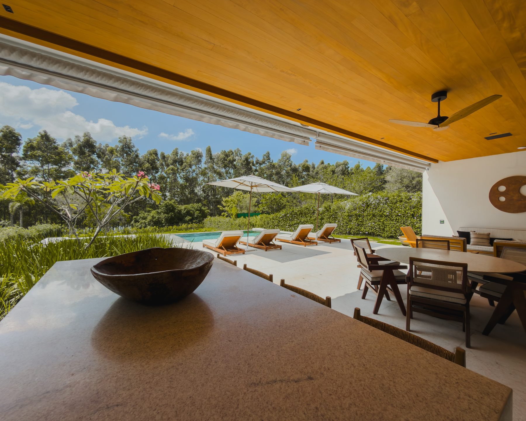 Bancada de pedra com gamela de madeira, com a piscina, os guarda-sóis e o jardim ao fundo