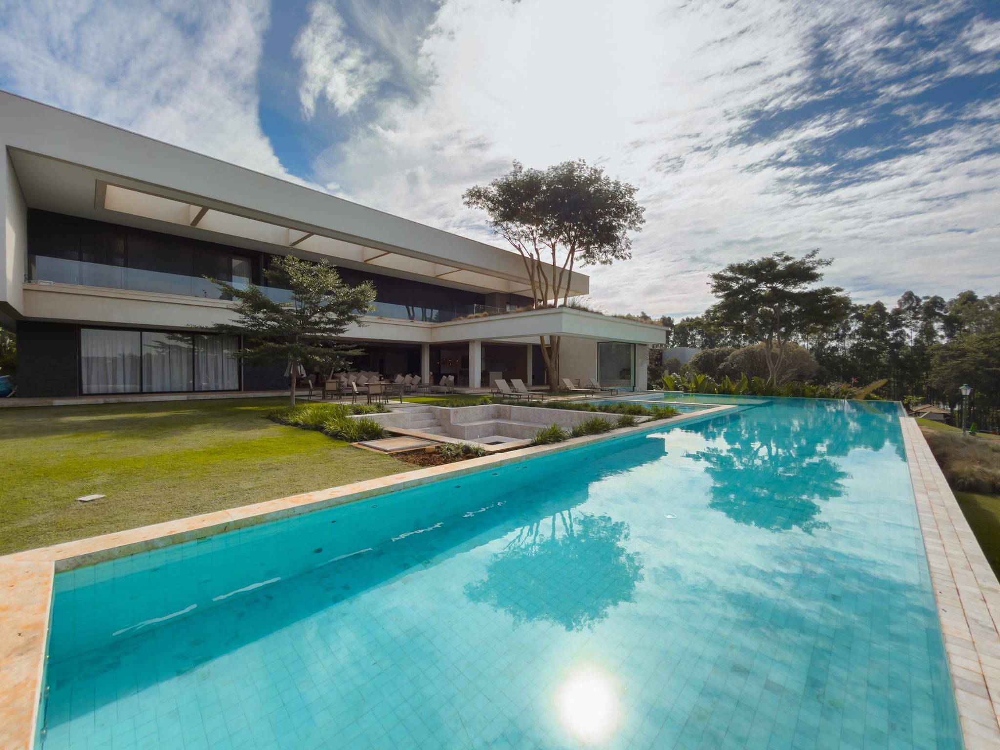 Casa moderna branca de dois pavimentos diante da piscina longa e do gramado, sob céu com nuvens