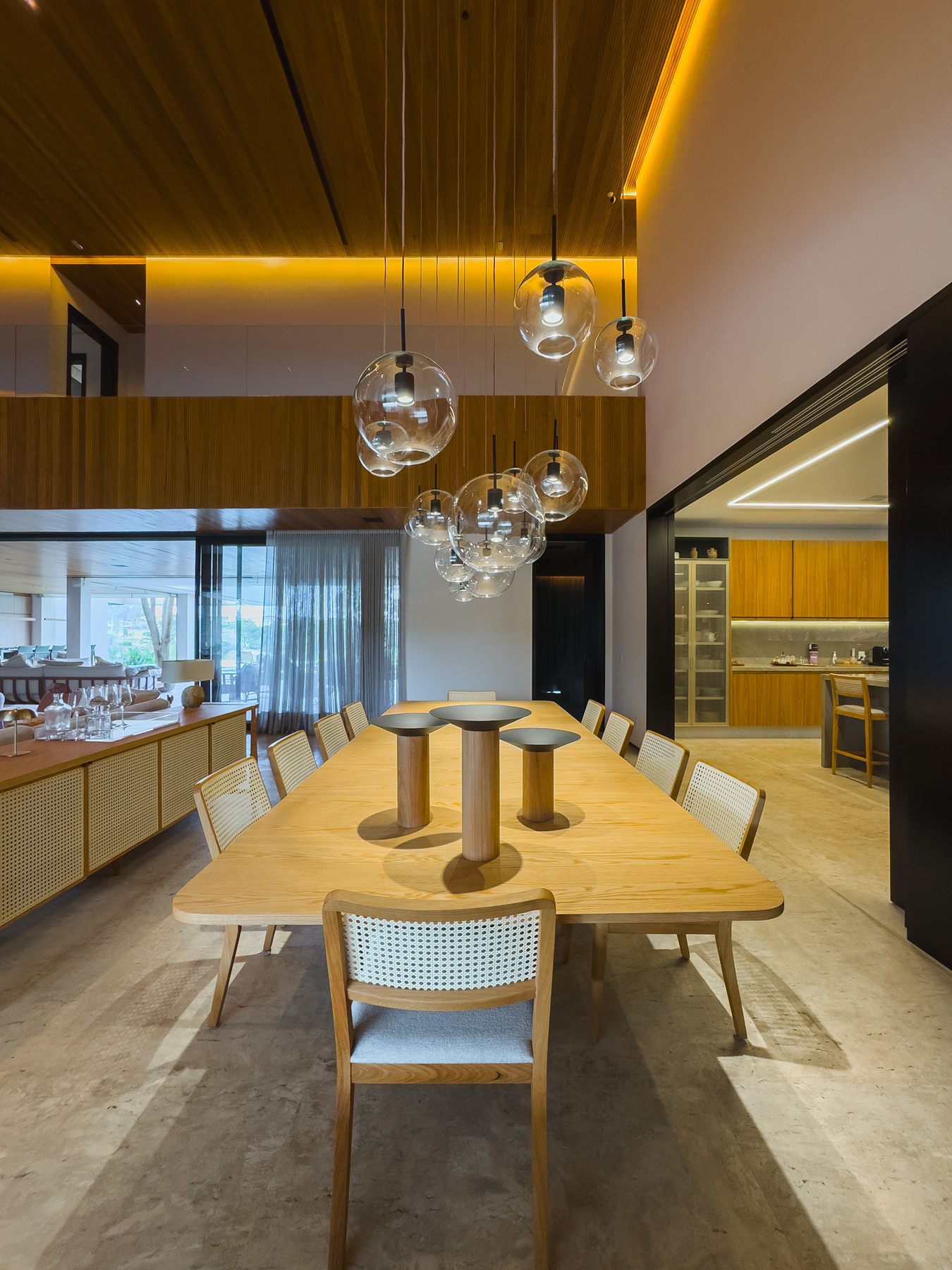 Mesa de jantar de madeira sob luminárias de vidro, com o estar e as janelas ao fundo