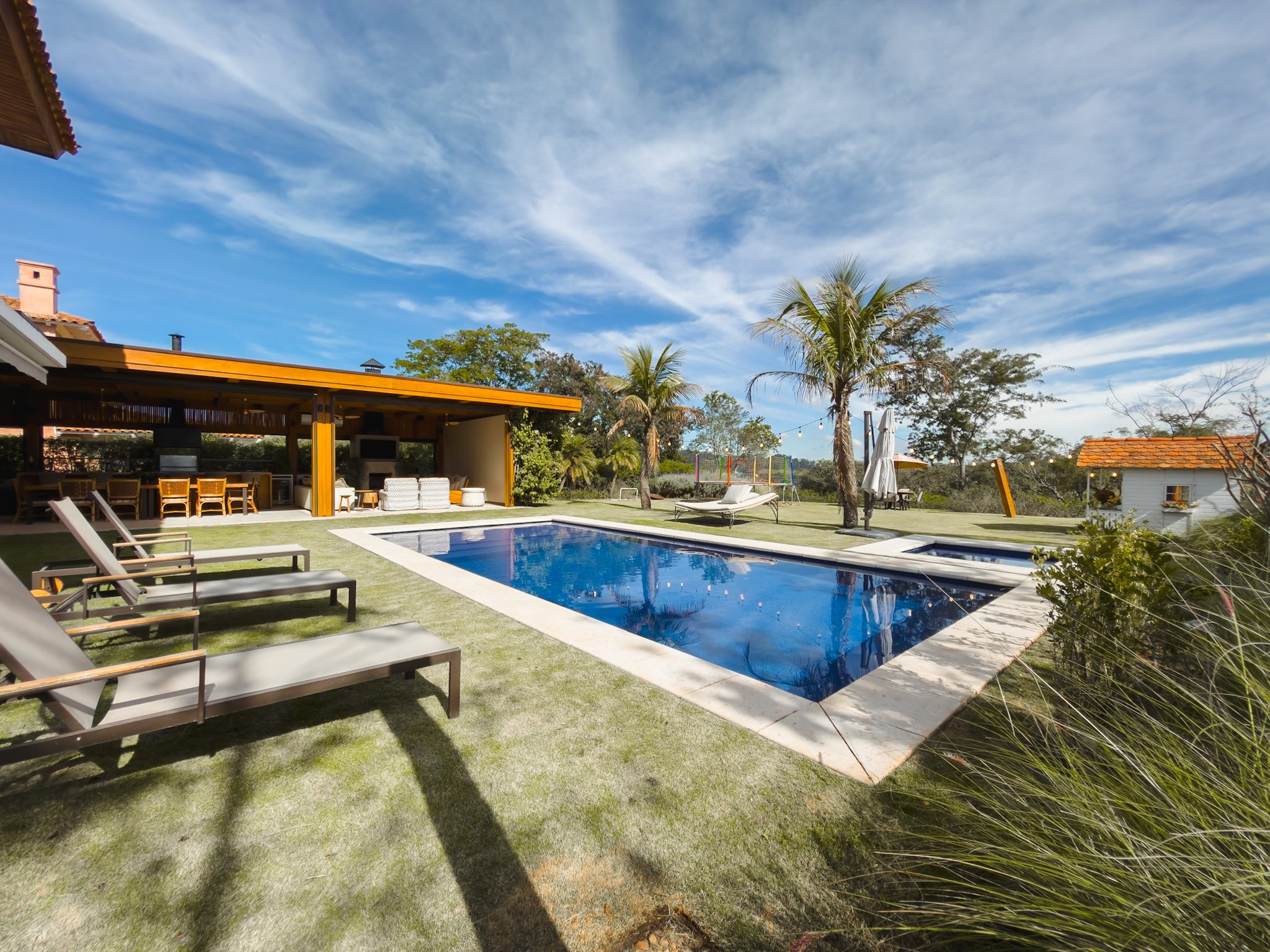 Piscina no gramado com espreguiçadeiras, o pergolado da área gourmet e palmeiras, sob céu com nuvens