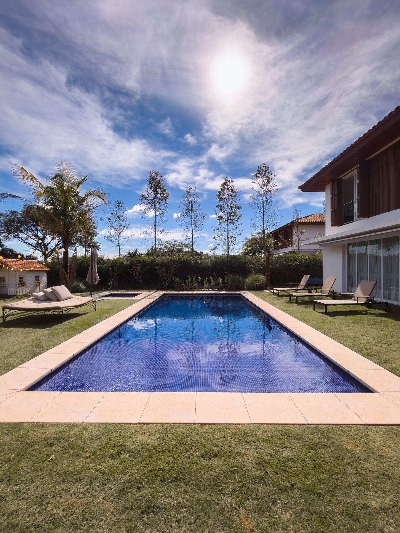 Piscina azul no gramado, com a casa de dois pavimentos ao lado e céu com nuvens