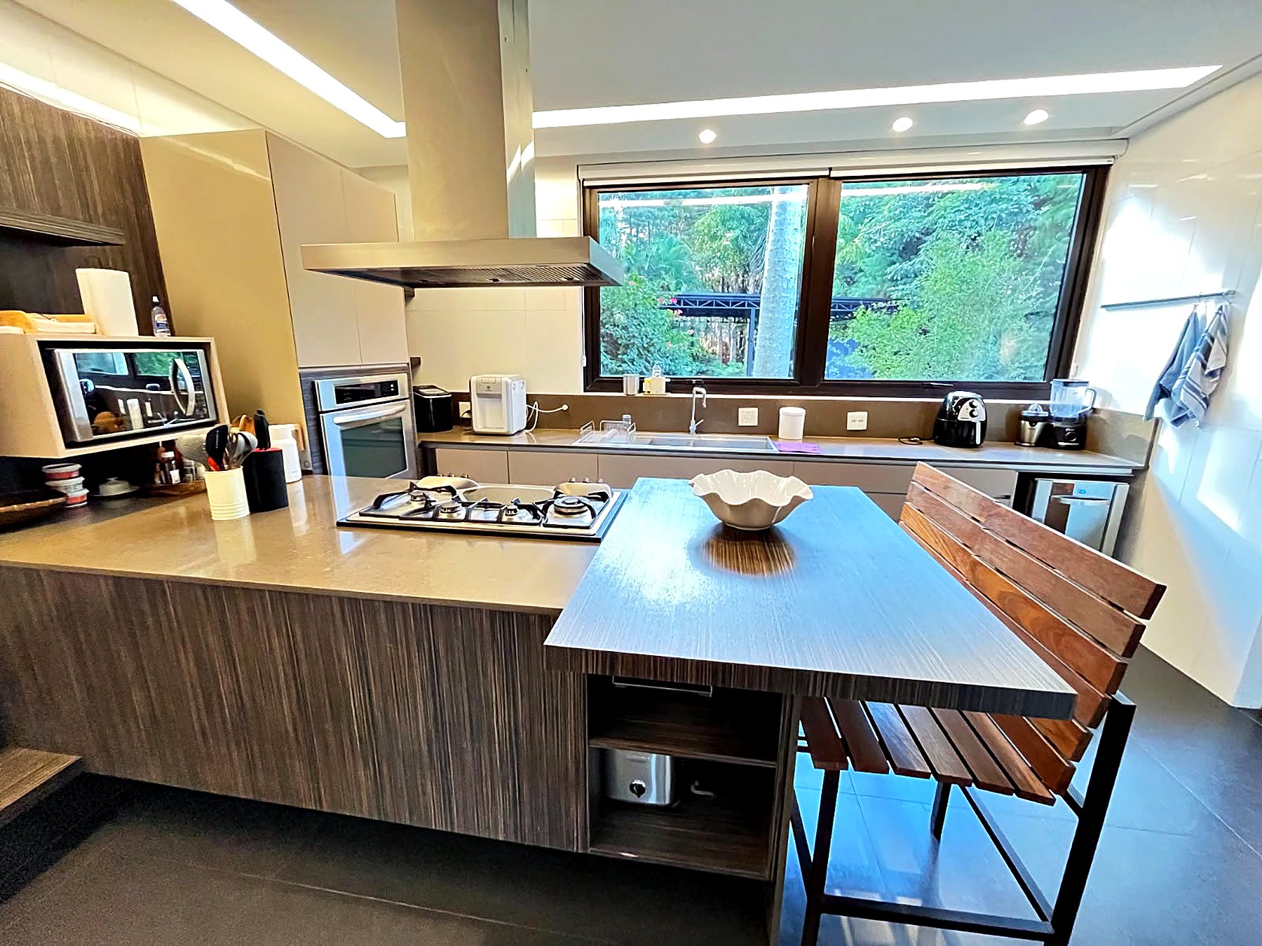 Cozinha com ilha de madeira, cooktop, coifa de inox e janela para as árvores
