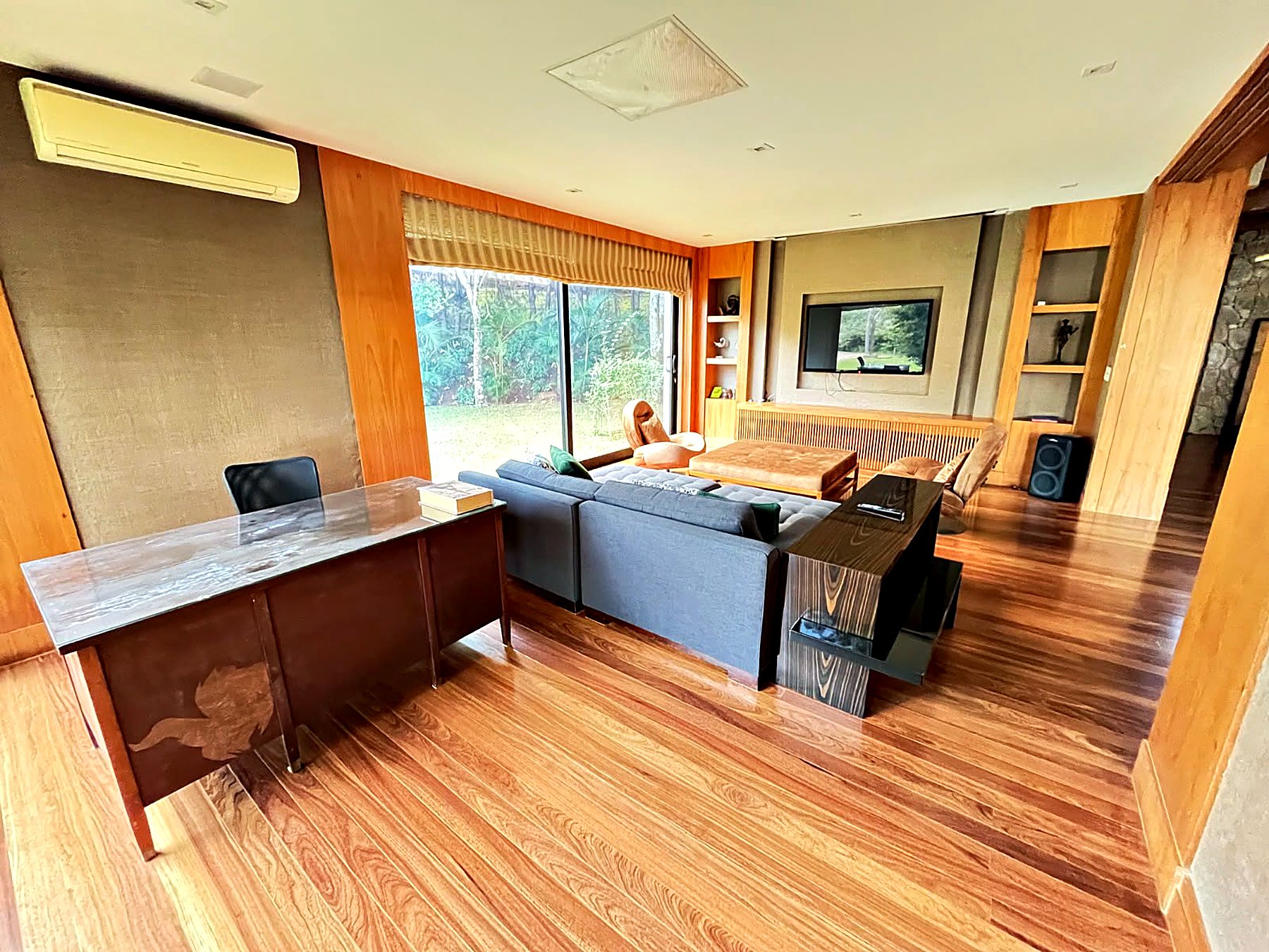 Escritório com mesa de madeira e pedra, sofá cinza e janela para o jardim