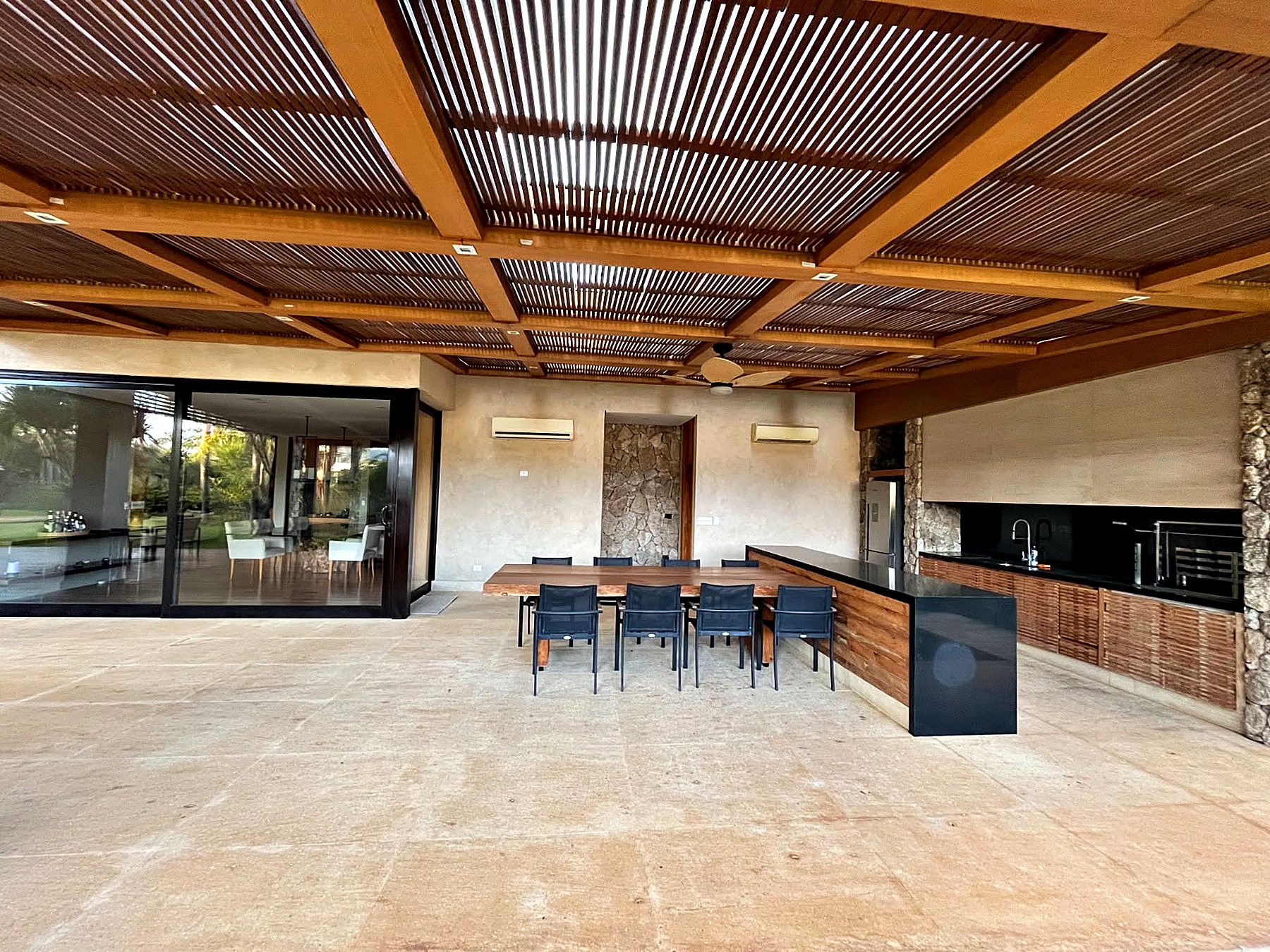 Espaço gourmet sob forro de madeira, com mesa longa, bancada escura e churrasqueira