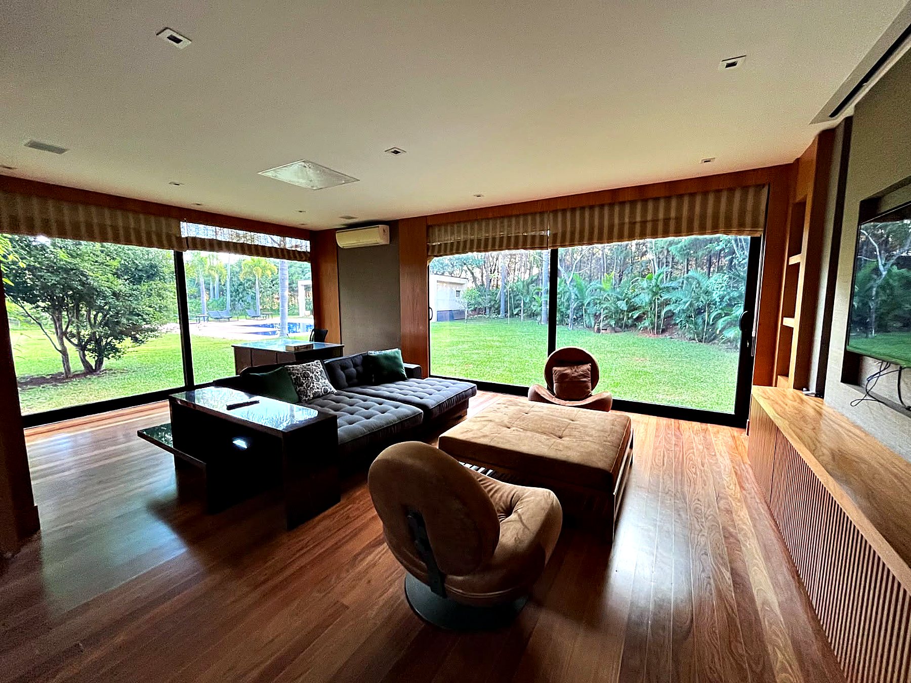 Sala com sofá escuro, poltrona de couro e janelas amplas para o gramado
