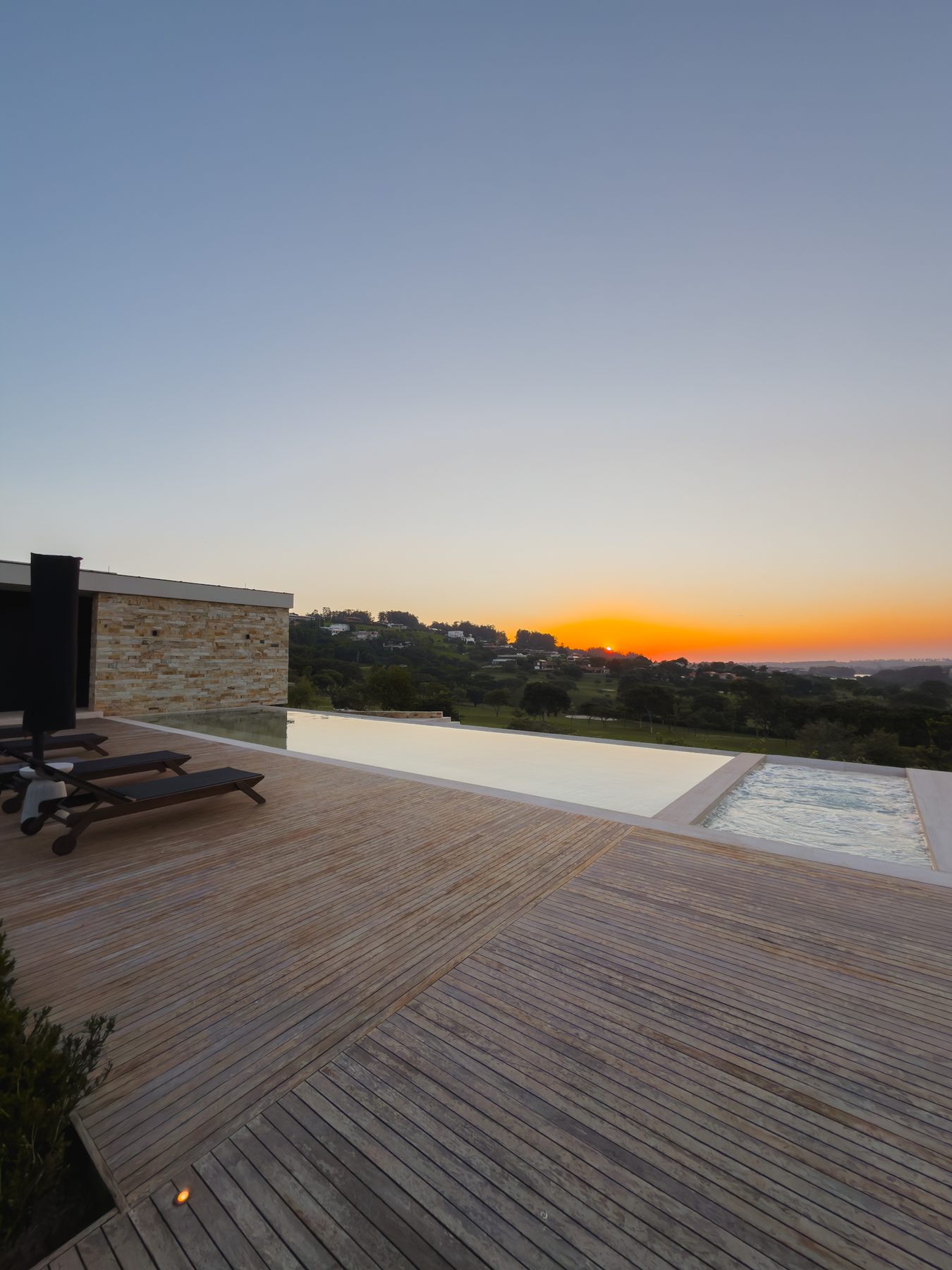 Deck de madeira junto à piscina ao pôr do sol, com o horizonte alaranjado