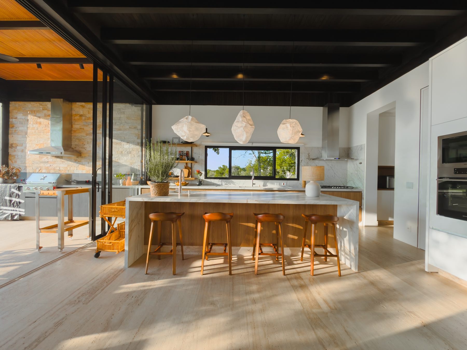 Cozinha com ilha branca, banquetas de madeira e luminárias pendentes, com janela para as árvores