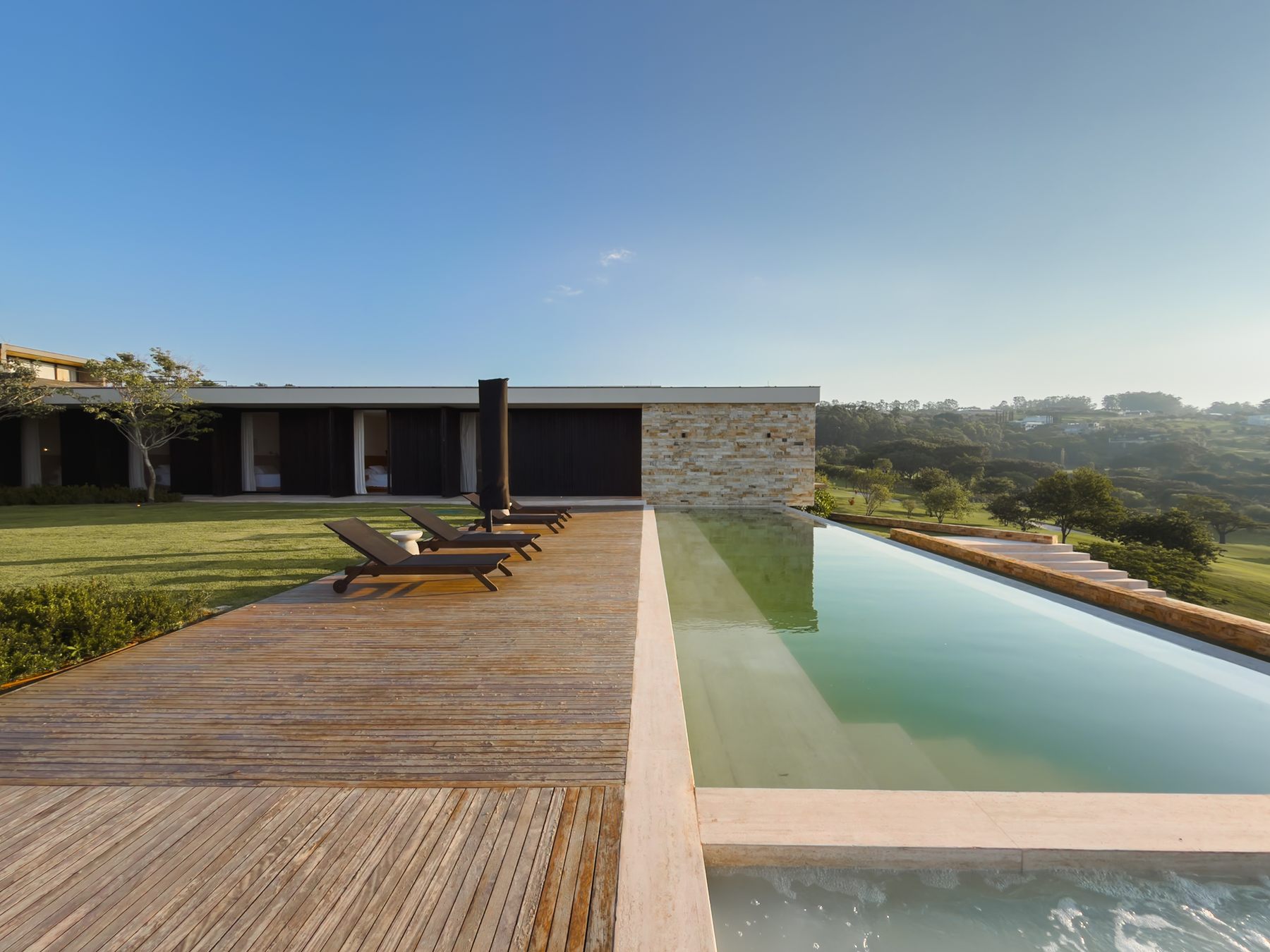 Deck de madeira com espreguiçadeiras ao lado da piscina, com a casa e o campo ao fundo