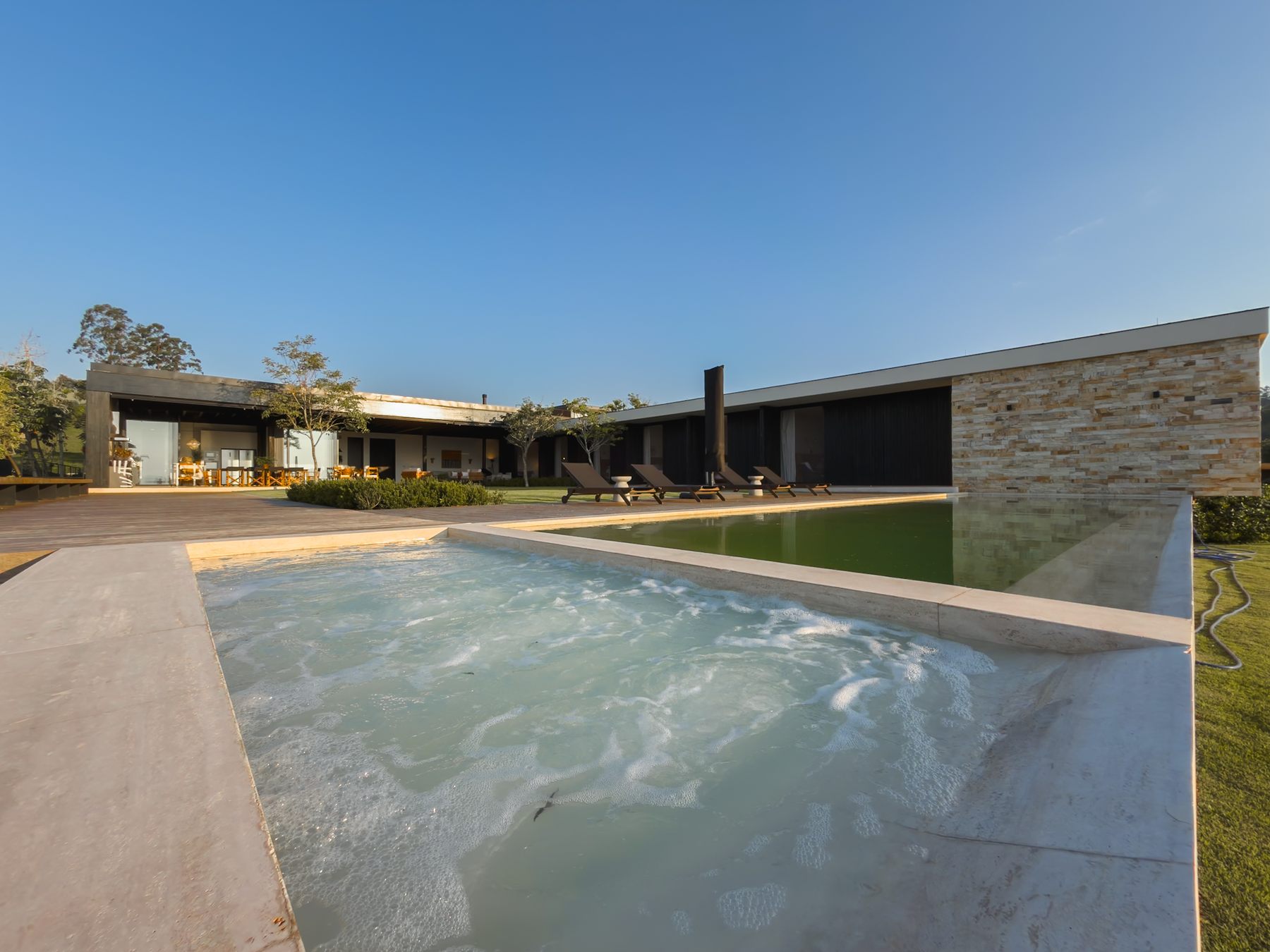 SPA com água em movimento ao lado da piscina, com a casa e o deck ao fundo sob céu azul