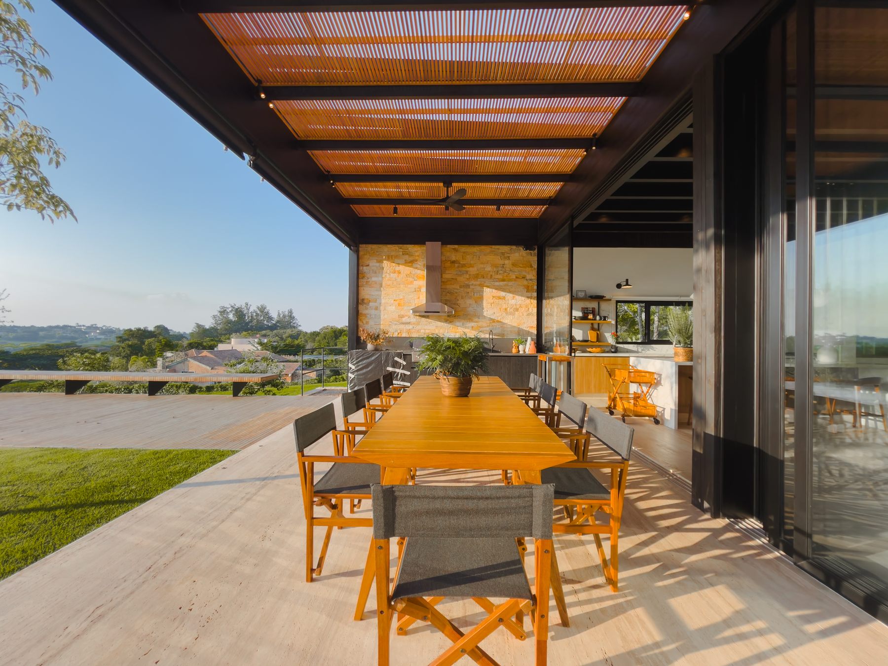 Varanda gourmet com mesa longa de madeira e cadeiras, sob pergolado, aberta para a vista