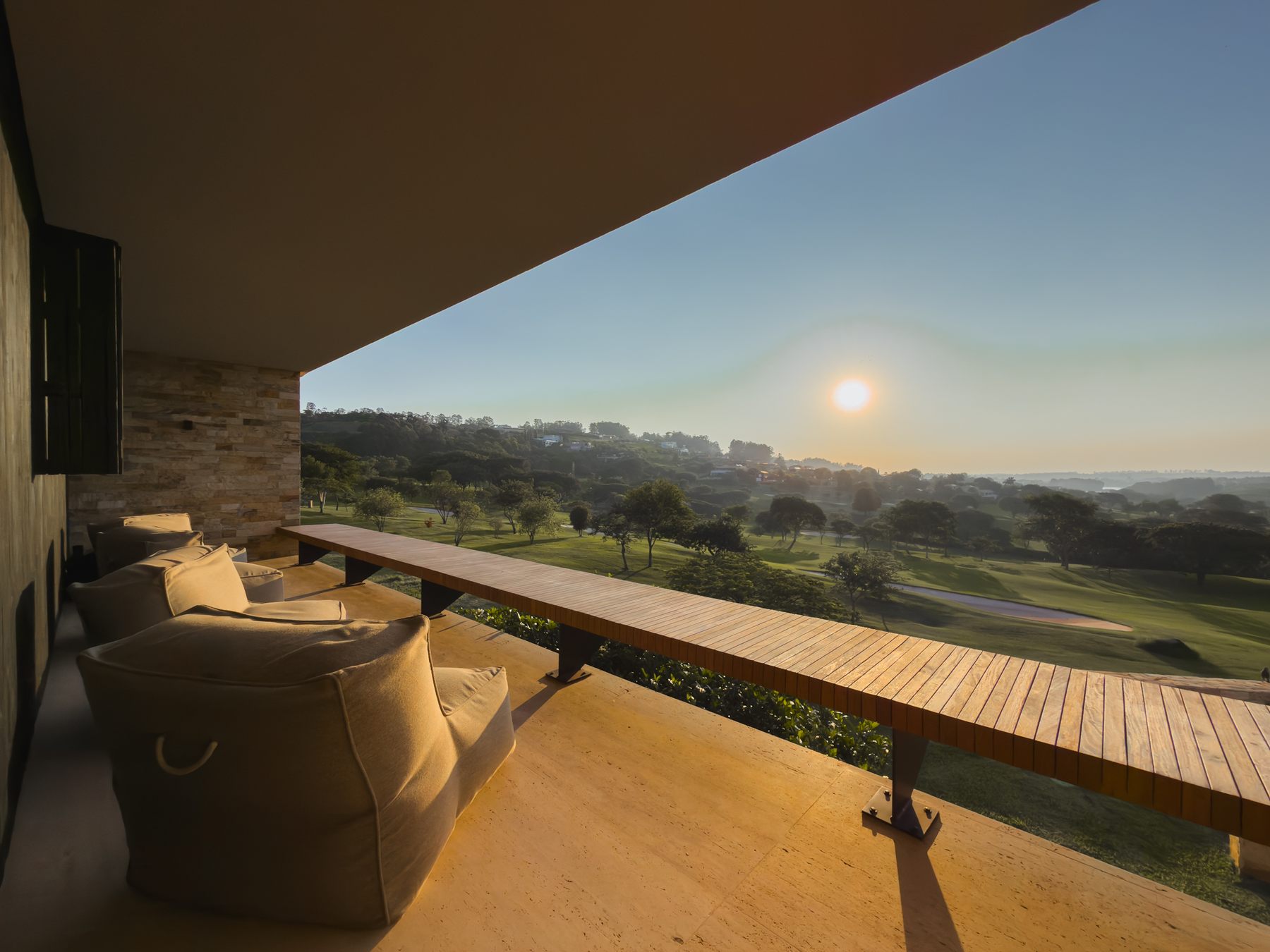 Varanda coberta com pufes e banco de madeira, diante do campo de golfe ao entardecer